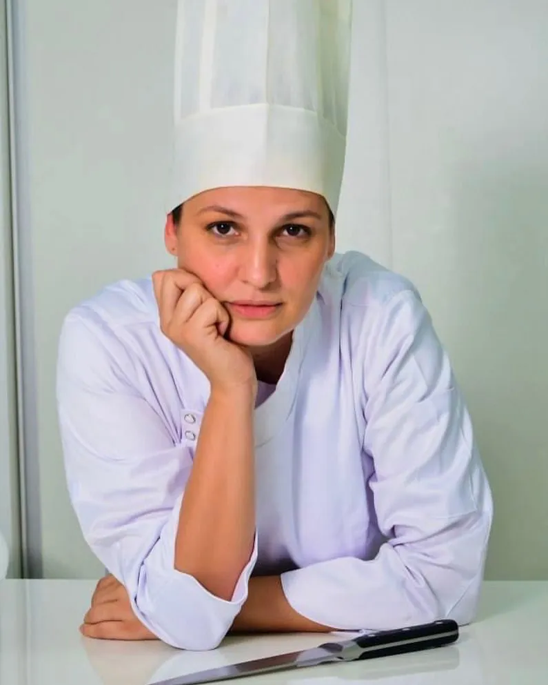 Retrato da chef Vanessa Bellini de dólmã branca e touca, apoiada na bancada
